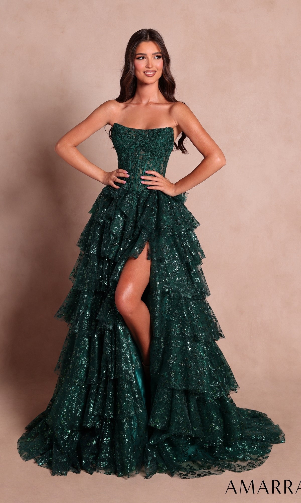 A woman wearing the Strapless Ruffled Prom Ball Gown: Amarra 89483 in emerald, featuring sequin tiered layers and a high slit, stands against a plain beige background.